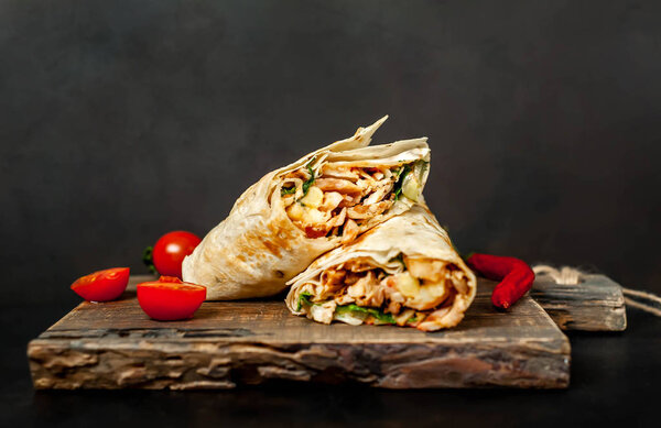 Traditional mexican burrito wrapped with chicken and vegetables served with ripe tomatoes on wooden chopping board 