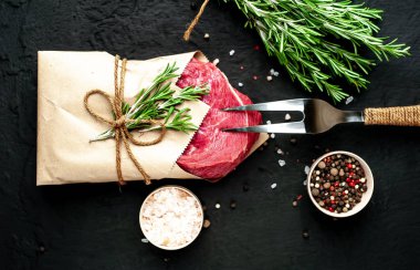 Çiğ sığır eti biftek taş arka plan üzerinde biberiye dalları ve baharatlar ile zanaat kağıt sarılmış 