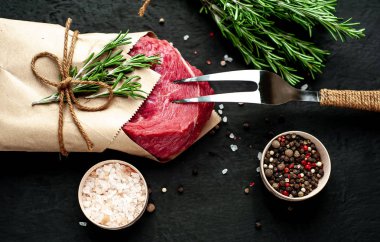 Çiğ sığır eti biftek taş arka plan üzerinde biberiye dalları ve baharatlar ile zanaat kağıt sarılmış 