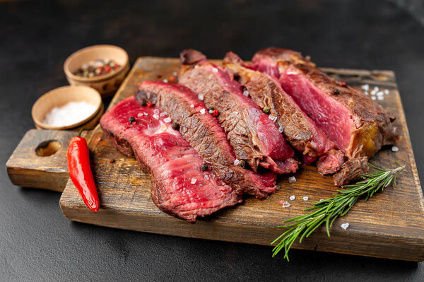 Fresh rare grilled beefsteak with spices on wooden chopping board over black stone background