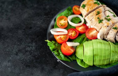 Izgara tavuk göğsü ve kiraz domatesli avokado salatası, ıspanak, taştan bir arka planda siyah bir tabak ve metin için fotokopi alanı.