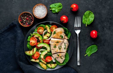 Izgara tavuk göğsü ve kiraz domatesli avokado salatası, ıspanak, taştan bir arka planda siyah bir tabak ve metin için fotokopi alanı.