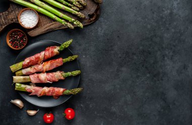 Pişmiş kuşkonmaz, domuz pastırması ve baharatla siyah bir tabakta, arka planda.