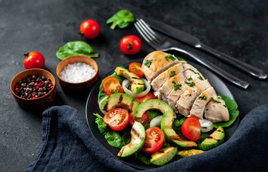 Domatesli tavuk, avokado dilimleri ve siyah tabakta yeşil salata. 