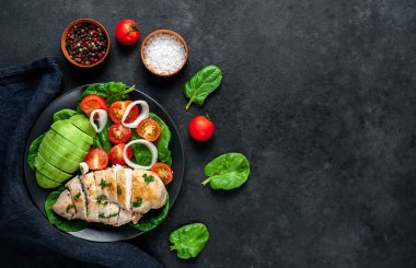 Domatesli tavuk, avokado dilimleri ve siyah tabakta yeşil salata. 