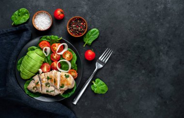 Domatesli tavuk, avokado dilimleri ve siyah tabakta yeşil salata. 