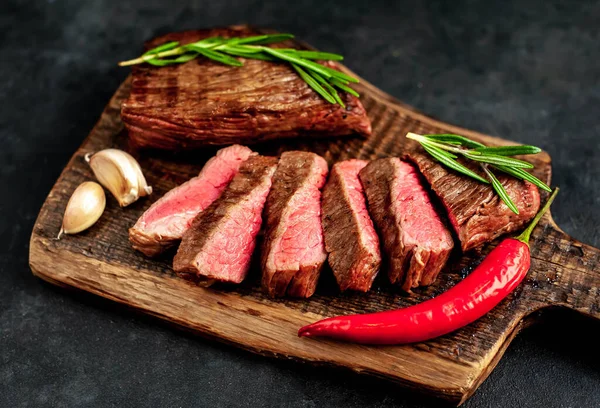 Fatty grilled beef steak with spices, rosemary and chili pepper on chopping board over black stone background