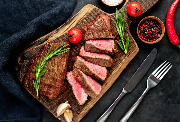 Fatty grilled beef steak with spices, rosemary and chili pepper on chopping board over black stone background