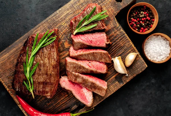 Fatty grilled beef steak with spices, rosemary and chili pepper on chopping board over black stone background