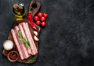 Baharatlı, sebzeli ve otlu çiğ sosisler bir şişe zeytinyağıyla ahşap kesme tahtasının üzerinde. Koyu arkaplan.