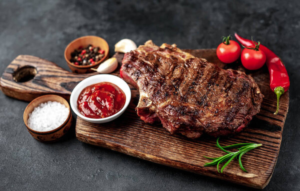 Grilled steak with spices, vegetables and herbs on a wooden cutting board with dark background.