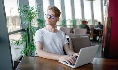 Beyaz tişörtlü ve gözlüklü genç adam kafeteryadaki bir masada dizüstü bilgisayarla çalışıyor.