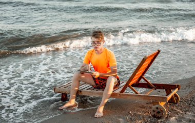 Tişörtlü ve şortlu bir adam tablet tutuyor, deniz kenarındaki ahşap iskemlede oturuyor.