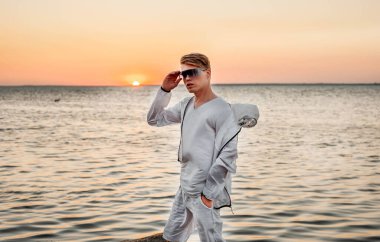 Beyaz elbiseli bir adam deniz kenarında gün batımına bakıyor.