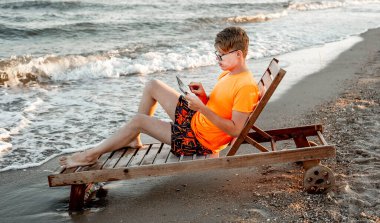 Tişörtlü ve şortlu bir adam tablet tutuyor, deniz kenarındaki ahşap iskemlede oturuyor.