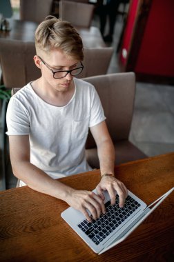 Beyaz tişörtlü ve gözlüklü genç adam kafeteryadaki bir masada dizüstü bilgisayarla çalışıyor.