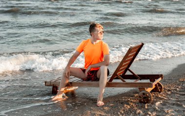 Tişörtlü ve şortlu bir adam deniz kenarındaki ahşap iskemlede oturuyor.