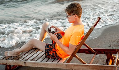 Tişörtlü ve şortlu bir adam tablet tutuyor, deniz kenarındaki ahşap iskemlede oturuyor.