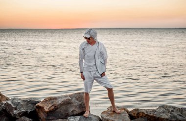 Beyaz elbiseli bir adam deniz kenarında gün batımına bakıyor.