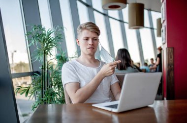 Beyaz tişörtlü ve gözlüklü genç adam kafeteryadaki bir masada dizüstü bilgisayarla çalışıyor.