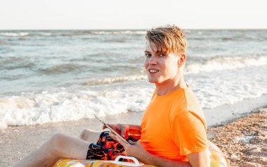 Tişörtlü ve şortlu bir adam tablet tutuyor, deniz kenarındaki ahşap iskemlede oturuyor.