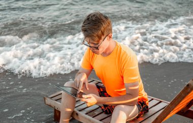 Tişörtlü ve şortlu bir adam tablet tutuyor, deniz kenarındaki ahşap iskemlede oturuyor.