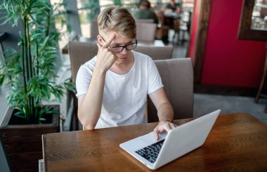 Beyaz tişörtlü ve gözlüklü genç adam kafeteryadaki bir masada dizüstü bilgisayarla çalışıyor.