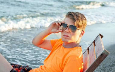 Tişörtlü ve şortlu bir adam deniz kenarındaki ahşap iskemlede oturuyor.