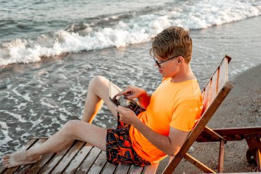 Tişörtlü ve şortlu bir adam tablet tutuyor, deniz kenarındaki ahşap iskemlede oturuyor.