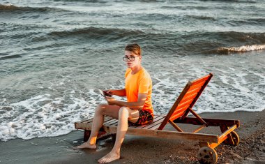 Tişörtlü ve şortlu bir adam tablet tutuyor, deniz kenarındaki ahşap iskemlede oturuyor.