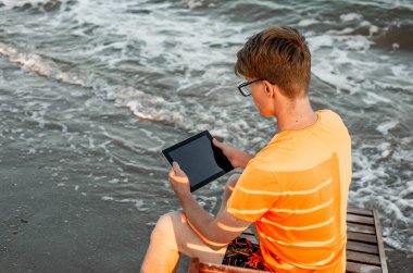 Tişörtlü ve şortlu bir adam tablet tutuyor, deniz kenarındaki ahşap iskemlede oturuyor.