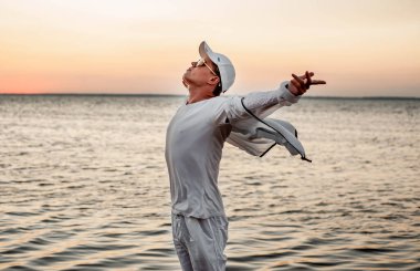 Beyaz elbiseli bir adam deniz kenarında gün batımına bakıyor.