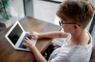 Beyaz tişörtlü ve gözlüklü genç adam kafeteryadaki bir masada dizüstü bilgisayarla çalışıyor.