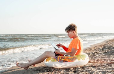 Tişörtlü ve şortlu bir adam tablet tutuyor, deniz kenarındaki ahşap iskemlede oturuyor.