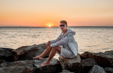 Beyaz elbiseli bir adam deniz kenarında gün batımına bakıyor.