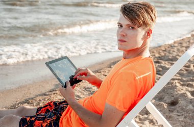 Tişörtlü ve şortlu bir adam tablet tutuyor, deniz kenarındaki ahşap iskemlede oturuyor.