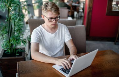 Beyaz tişörtlü ve gözlüklü genç adam kafeteryadaki bir masada dizüstü bilgisayarla çalışıyor.