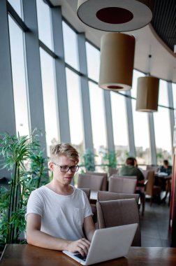 Beyaz tişörtlü ve gözlüklü genç adam kafeteryadaki bir masada dizüstü bilgisayarla çalışıyor.