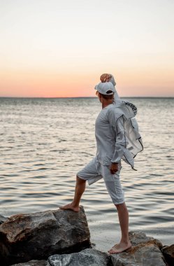 Beyaz elbiseli bir adam deniz kenarında gün batımına bakıyor.