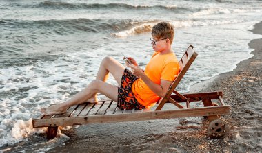 Tişörtlü ve şortlu bir adam tablet tutuyor, deniz kenarındaki ahşap iskemlede oturuyor.