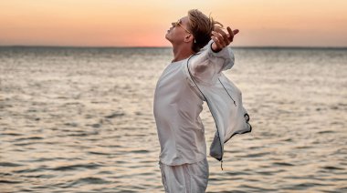 Beyaz elbiseli bir adam deniz kenarında gün batımına bakıyor.