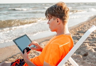 Tişörtlü ve şortlu bir adam tablet tutuyor, deniz kenarındaki ahşap iskemlede oturuyor.