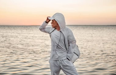 Beyaz elbiseli bir adam deniz kenarında gün batımına bakıyor.