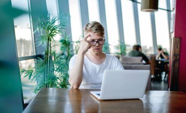 Beyaz tişörtlü ve gözlüklü genç adam kafeteryadaki bir masada dizüstü bilgisayarla çalışıyor.