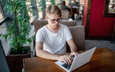 Beyaz tişörtlü ve gözlüklü genç adam kafeteryadaki bir masada dizüstü bilgisayarla çalışıyor.