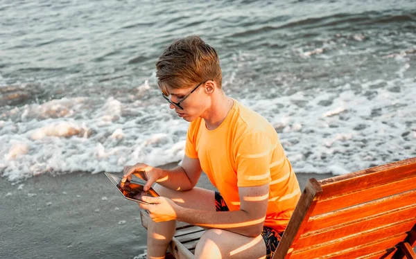 Tişörtlü ve şortlu bir adam tablet tutuyor, deniz kenarındaki ahşap iskemlede oturuyor.