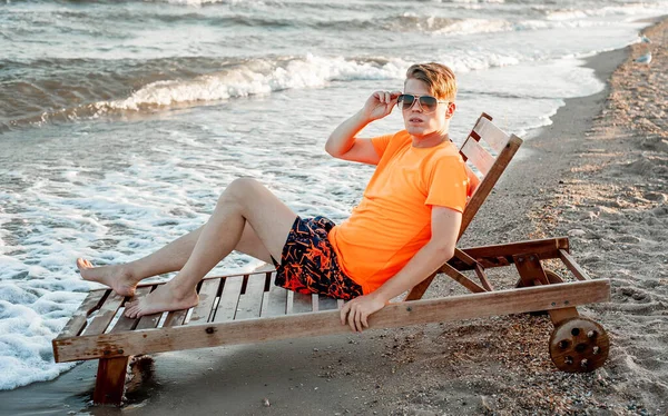 Tişörtlü ve şortlu bir adam deniz kenarındaki ahşap iskemlede oturuyor.