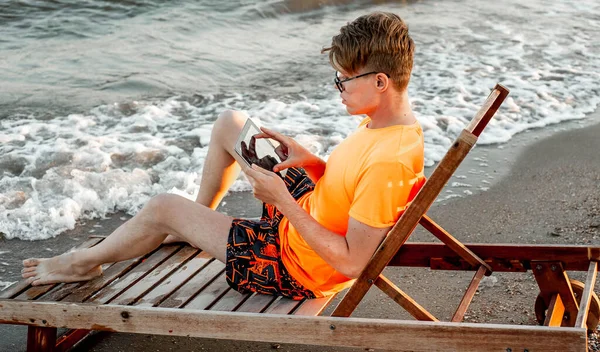 Tişörtlü ve şortlu bir adam tablet tutuyor, deniz kenarındaki ahşap iskemlede oturuyor.