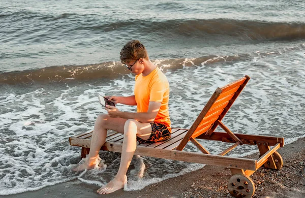Tişörtlü ve şortlu bir adam tablet tutuyor, deniz kenarındaki ahşap iskemlede oturuyor.