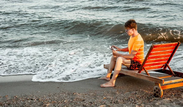 Tişörtlü ve şortlu bir adam tablet tutuyor, deniz kenarındaki ahşap iskemlede oturuyor.